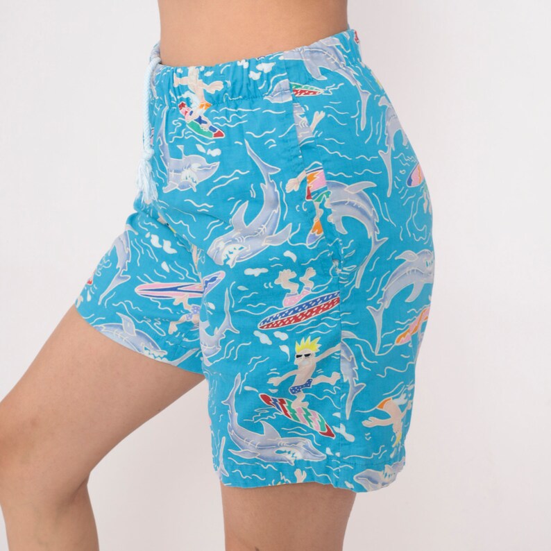 K&ouml;nnte beinhalten: Blaue Badeshorts mit einem Muster aus Cartoon-Surfern und Haien. Die Shorts haben einen Kordelzug.