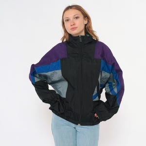 May include: A black, purple, blue, and gray vintage windbreaker jacket with a full zipper. The jacket has a high collar and elastic cuffs. The sleeves are accented with blue and gray panels. The jacket is worn by a person.