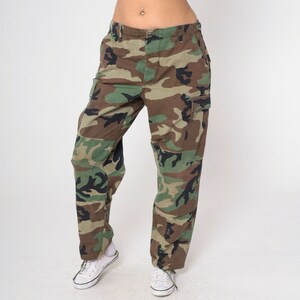 May include: Camouflage cargo pants in shades of green, brown, and black. The pants have multiple pockets and a classic military-inspired design. The pants are paired with white sneakers.