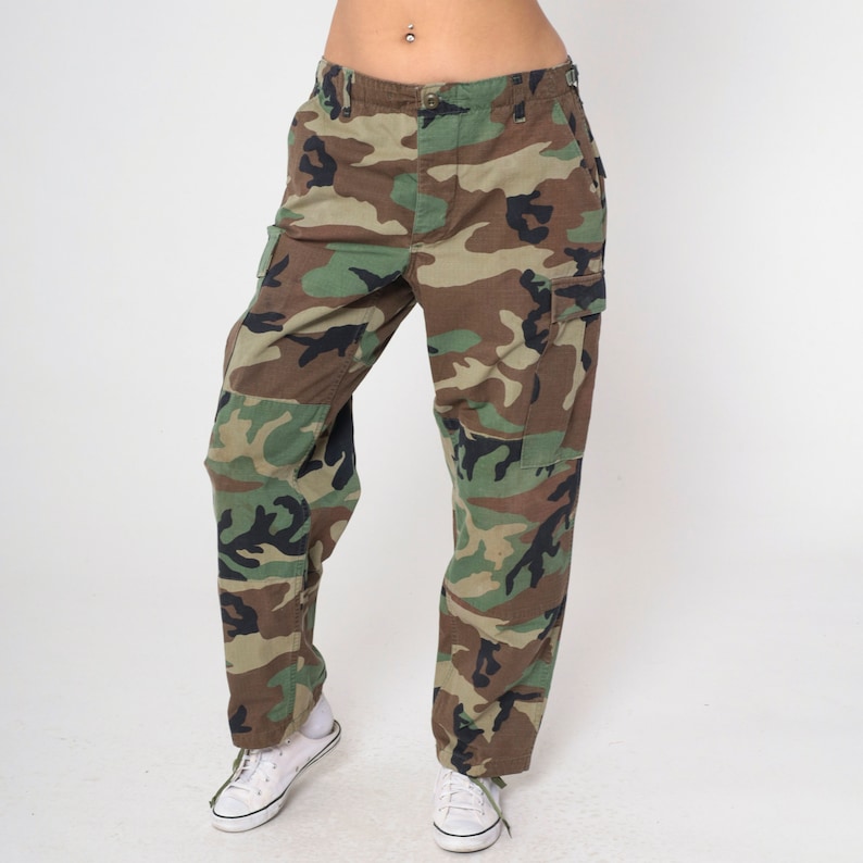 May include: Camouflage cargo pants in shades of green, brown, and black. The pants have multiple pockets and a classic military-inspired design. The pants are paired with white sneakers.