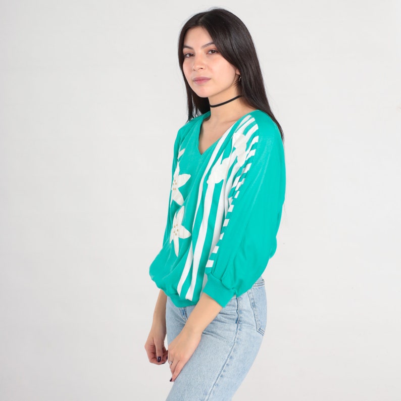 May include: A turquoise blue long-sleeved shirt with white stripes and white flower designs. The shirt has a v-neck and a relaxed fit.