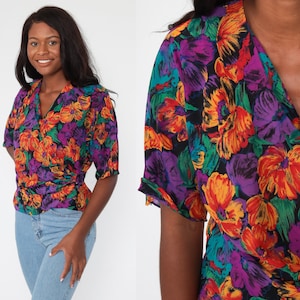 May include: A short-sleeved wrap top with a vibrant floral pattern in shades of orange, purple, and green on a black background. The top has a V-neck and a gathered waist. The model is wearing blue jeans.