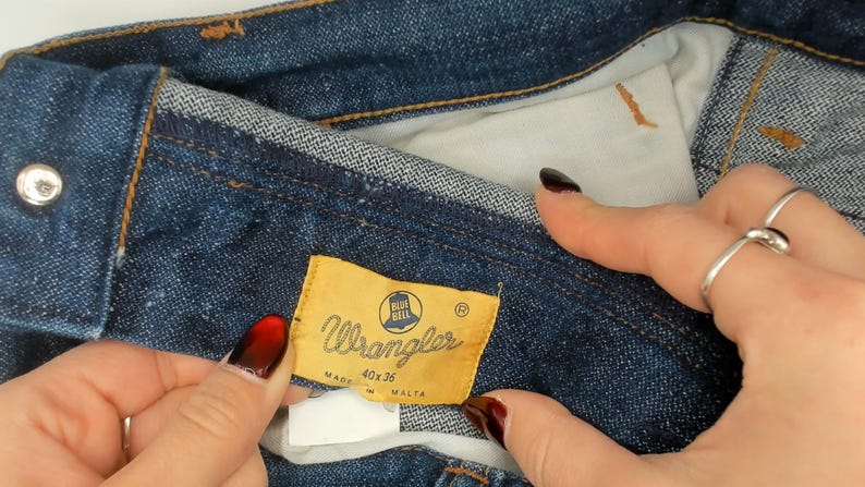 May include: Close-up of blue denim jeans with a Wrangler label. The label is yellow with the Wrangler logo and size information. The jeans are made in Malta. The size is 40x36.