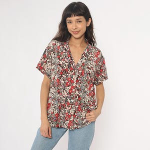 May include: Short-sleeved floral print shirt with a V-neck collar. The shirt features a pattern of red, white, and gray flowers on a black background. The shirt is buttoned down the front and is paired with light blue jeans.