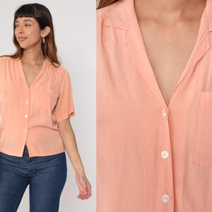 Puede incluir: Camisa melocot&oacute;n de manga corta con cuello y bolsillo en el pecho. La camisa est&aacute; abotonada. La modelo lleva vaqueros azules. La camisa tiene un corte relajado.