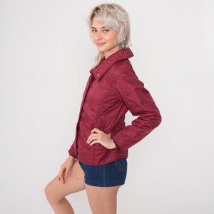 May include: A burgundy quilted jacket with a button-up front and a wide collar. The jacket has a fitted silhouette and is paired with denim shorts. The jacket is a solid color and appears to be made of a lightweight material.