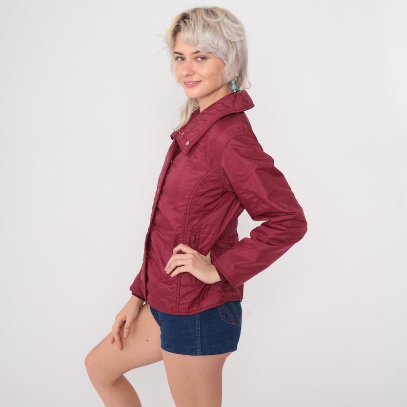 May include: A burgundy quilted jacket with a button-up front and a wide collar. The jacket has a fitted silhouette and is paired with denim shorts. The jacket is a solid color and appears to be made of a lightweight material.