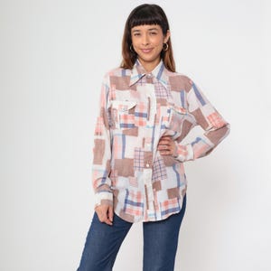 May include: A patchwork button-down shirt with a collared neckline. The shirt features a mix of brown, white, pink, and blue plaid and solid patches. It has two chest pockets and long sleeves. The model is wearing blue jeans.