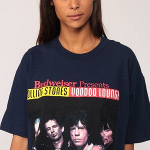 May include: Navy blue t-shirt featuring a graphic of The Rolling Stones with the text "Budweiser Presents Rolling Stones Voodoo Lounge" in red, yellow, and white. The shirt has short sleeves and a crew neck.
