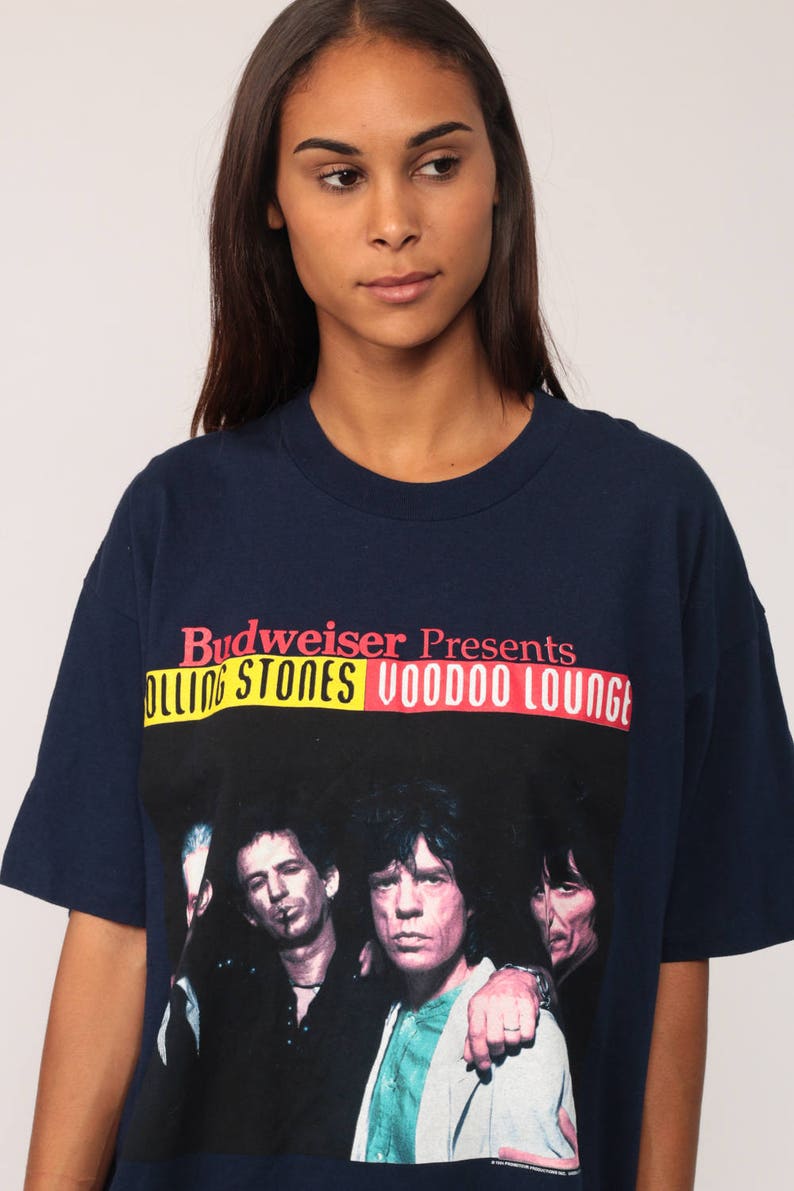 May include: Navy blue t-shirt featuring a graphic of The Rolling Stones with the text "Budweiser Presents Rolling Stones Voodoo Lounge" in red, yellow, and white. The shirt has short sleeves and a crew neck.