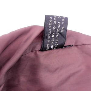 May include: Close-up of a burgundy-colored garment, possibly a jacket or coat, with a black tag attached. The tag displays text including "OUT SHELL: GENUINE", "LINING: NYLON 100%", and "PROFESSIONAL D".