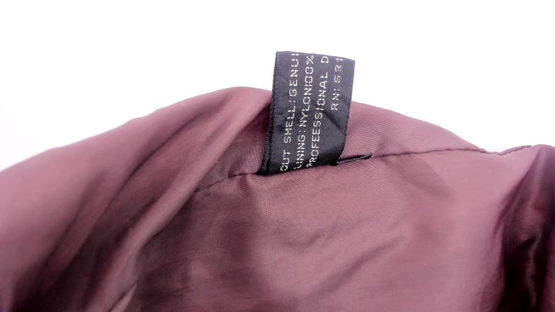 May include: Close-up of a burgundy-colored garment, possibly a jacket or coat, with a black tag attached. The tag displays text including "OUT SHELL: GENUINE", "LINING: NYLON 100%", and "PROFESSIONAL D".