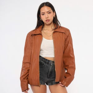 May include: A brown leather jacket with a zipper closure. The jacket has a collared neckline and long sleeves. The model is wearing a white top and denim shorts. The jacket is a medium brown color.