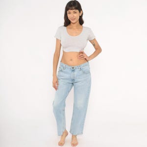 May include: A light gray, short-sleeved crop top with a scoop neck and light blue jeans. The jeans have a straight leg fit. The person is standing in front of a white background.