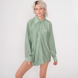 May include: A long-sleeved, button-down shirt in a soft, muted green color. The shirt has a classic collar, a chest pocket, and buttoned cuffs. The fabric appears to be a lightweight material, suitable for warmer weather. The shirt is unbuttoned and worn as a top.