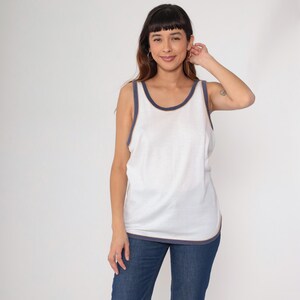 May include: White tank top with navy blue trim. The sleeveless top has a rounded neckline and a relaxed fit. The trim is visible around the neckline and armholes, as well as the bottom hem. The model is wearing blue jeans.