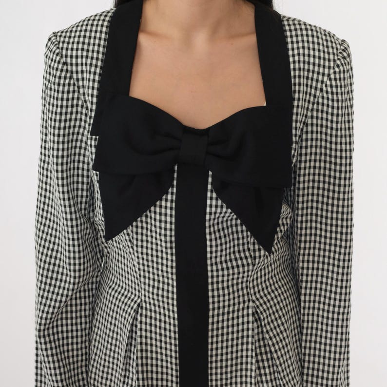 May include: A black and white gingham dress with a large black bow at the neckline. The dress has long sleeves and a fitted bodice.