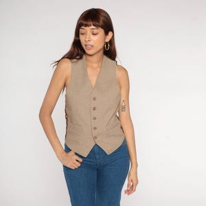 May include: A tan vest with a V-neck and button closure. The vest has a tailored fit and is worn with blue jeans. The vest is sleeveless and has a subtle texture. The back of the vest has a patterned design.