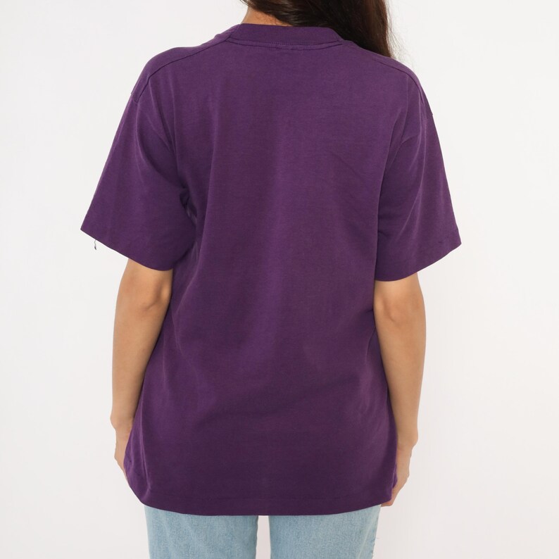 May include: A solid purple t-shirt with short sleeves. The shirt is a deep shade of purple and appears to be made of a soft material. The back of the shirt is visible, and it is worn with light blue jeans.