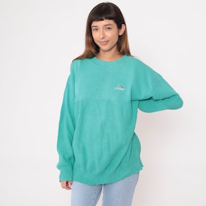 Puede incluir: Un suéter de color turquesa con cuello redondo y mangas largas. Las mangas presentan un diseño de punto texturizado y el pecho un panel liso. Un pequeño logotipo de Jaguar está bordado en el pecho. El modelo lleva vaqueros azul claro.