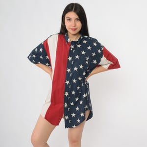 May include: Button-down shirt featuring a patriotic design. The shirt is divided into sections with red and white stripes on one side and a navy blue field of white stars on the other. The shirt has short sleeves.