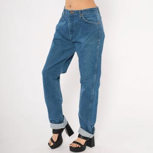 May include: Blue denim jeans with a button closure and front pockets. The jeans have a straight leg fit and are cuffed at the ankles. The model is wearing black platform heels.