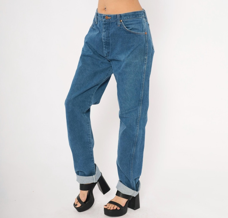 May include: Blue denim jeans with a button closure and front pockets. The jeans have a straight leg fit and are cuffed at the ankles. The model is wearing black platform heels.