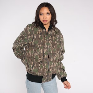 May include: A camouflage bomber jacket with a black collar, cuffs, and hem. The jacket features a full-length zipper and a pattern of green leaves and brown bark-like shapes. The model is wearing light blue jeans.