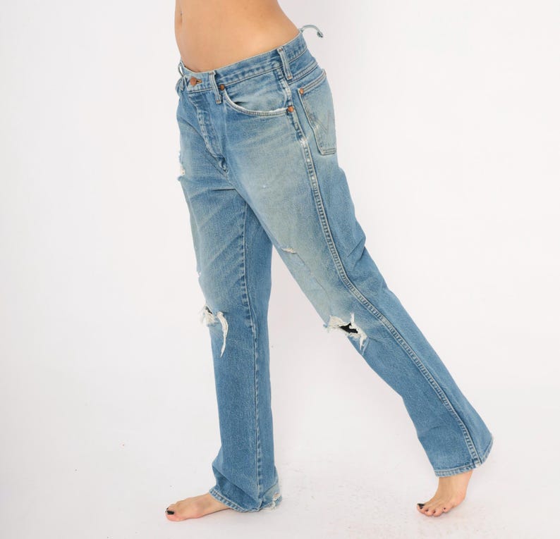 May include: Light blue denim jeans with distressed details, including rips at the knees. The jeans have a classic five-pocket design and a faded wash. The jeans are a straight-leg style.