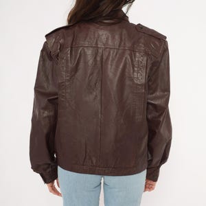 May include: A brown leather jacket with a classic bomber style. The jacket features a stand-up collar, shoulder epaulets, and a zip-up front. The sleeves are long and end in a ribbed cuff. The jacket is worn over blue jeans.