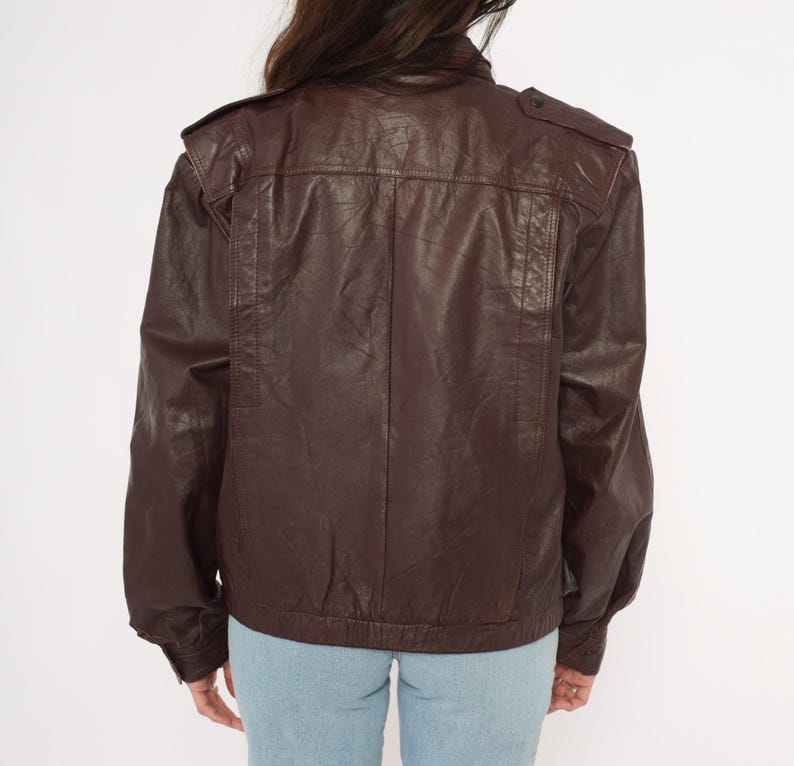 May include: A brown leather jacket with a classic bomber style. The jacket features a stand-up collar, shoulder epaulets, and a zip-up front. The sleeves are long and end in a ribbed cuff. The jacket is worn over blue jeans.