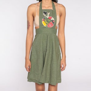 May include: Olive green apron dress with an apple applique. The apple design features a patchwork of colorful fabrics. The dress has a halter neckline, a fitted bodice, and a flared skirt. The apron has pockets and a cut-out design at the waist.