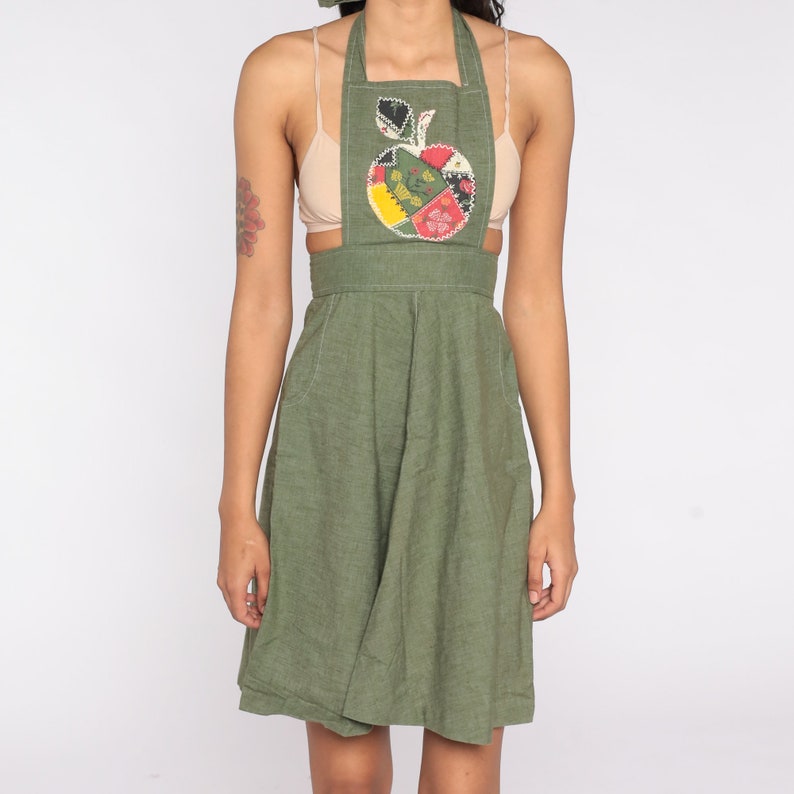 May include: Olive green apron dress with an apple applique. The apple design features a patchwork of colorful fabrics. The dress has a halter neckline, a fitted bodice, and a flared skirt. The apron has pockets and a cut-out design at the waist.