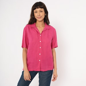 May include: A short-sleeved, hot pink button-down shirt with a collar. The shirt is worn with blue jeans. The shirt has white buttons down the front. The model is smiling.