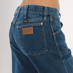 May include: Close-up of blue denim jeans with a brown leather patch on the back pocket. The patch has the brand name "Wrangler" embossed on it. The jeans have copper rivets and a classic five-pocket design.