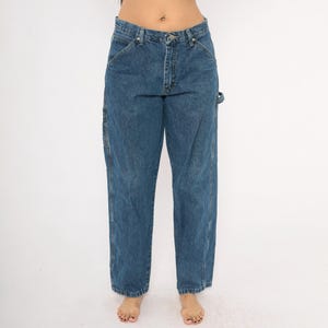 May include: Blue denim work pants with a classic five-pocket design. The pants have a straight leg cut and feature reinforced stitching and a utility pocket on the side. The jeans are a medium wash and have a button and zipper closure.