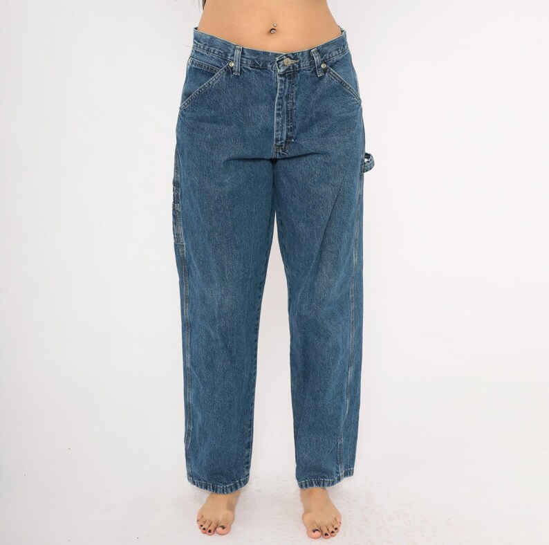 May include: Blue denim work pants with a classic five-pocket design. The pants have a straight leg cut and feature reinforced stitching and a utility pocket on the side. The jeans are a medium wash and have a button and zipper closure.