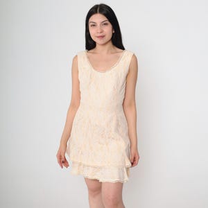 Puede incluir: Un vestido de encaje sin mangas de color crema con un ribete de perlas alrededor del escote y el dobladillo. El vestido tiene un corpiño ajustado y una falda acampanada.