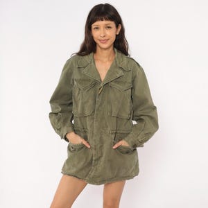 May include: An olive green field jacket featuring a collared neckline, long sleeves with buttoned cuffs, and four front pockets with flap closures. The jacket has a button-up front, a cinched waist detail, and a structured, utilitarian design.