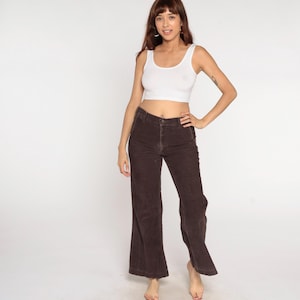 May include: A woman wearing a white tank top and brown corduroy pants. The pants have a flared leg design. The outfit is casual and suitable for everyday wear. The background is plain white.