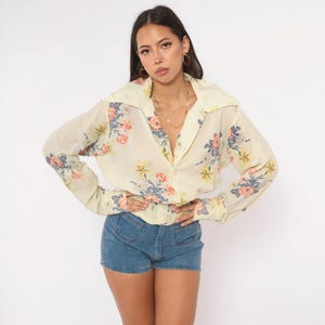 May include: A long-sleeve button-down shirt in a cream color featuring a floral pattern with orange, yellow, and blue flowers. The shirt has a wide collar, long sleeves with buttoned cuffs, and a gathered, elasticized waistline at the hem.