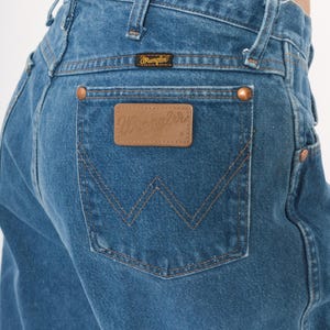 May include: Close-up of blue denim jeans with the Wrangler logo visible on the back pocket and waistband. The jeans have copper-colored rivets and stitching details. The back pocket features a distinctive W-shaped design.