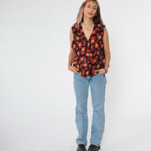 Puede incluir: Un chaleco negro sin mangas con estampado floral rojo y naranja. El chaleco tiene cuello en V y cierre de botones. La modelo lleva pantalones vaqueros azules.