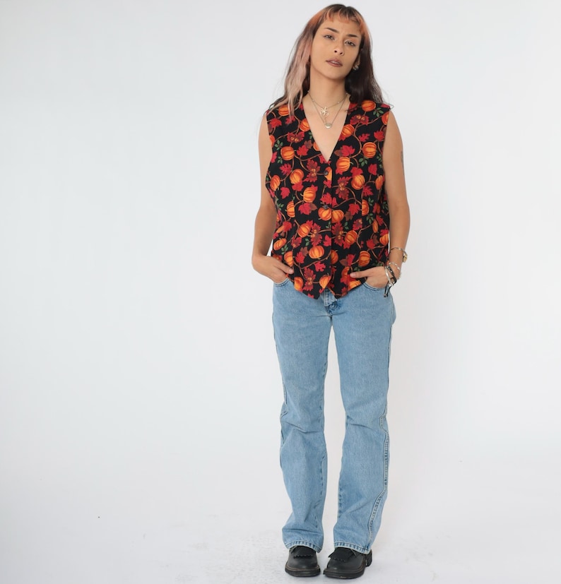 Puede incluir: Un chaleco negro sin mangas con estampado floral rojo y naranja. El chaleco tiene cuello en V y cierre de botones. La modelo lleva pantalones vaqueros azules.