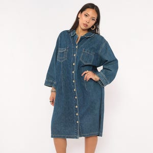 May include: A knee-length dark blue denim shirt dress featuring a classic collar, long sleeves with cuffs, and a full-length front button closure. The garment includes two chest pockets with flaps and vertical seam detailing along the front bodice.