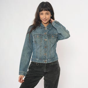 May include: A light blue denim jacket with a button-up front, two chest pockets, and long sleeves. The jacket has a classic design and is made of a durable denim material. The jacket is worn with dark gray pants.