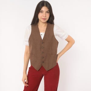 Peut inclure: Un gilet marron doté d'une encolure en V, de cinq boutons sur le devant et de deux petites poches passepoilées. Le gilet est porté sur une chemise blanche à manches courtes avec des détails en dentelle, associée à un pantalon rouge uni.
