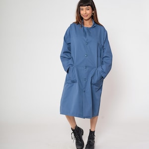 Peut inclure: Un long manteau de laboratoire bleu, boutonné, avec des poches. Le manteau arrive au genou et a des manches longues. La personne sur l'image porte des bottes noires.