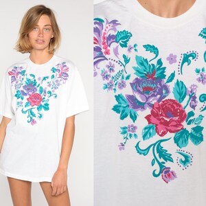 May include: White t-shirt with a floral print in shades of purple, pink, and teal. The flowers are arranged in a heart shape.
