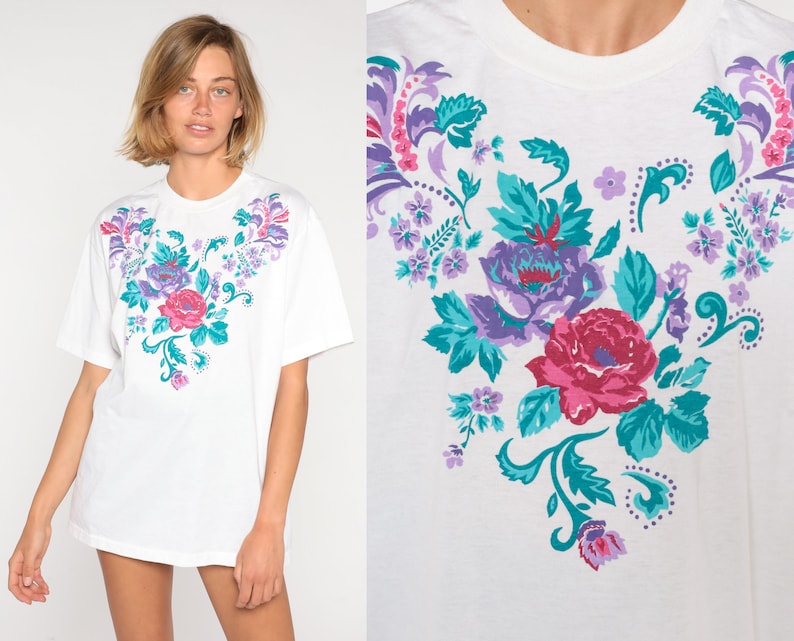 May include: White t-shirt with a floral print in shades of purple, pink, and teal. The flowers are arranged in a heart shape.